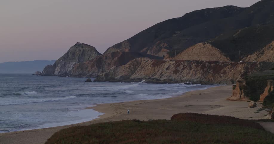 A person walking on an empty beach in the late afternoon near a promontory
