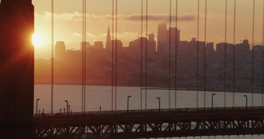 Traffic moves on a suspension bridge as the sun glows on the horizon behind the metropolitan city skyline