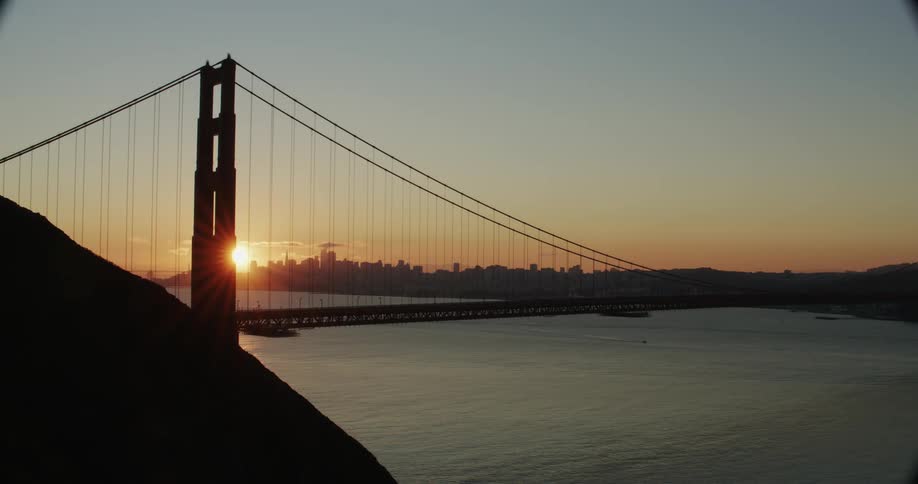 Traffic moves on a suspension bridge over the river as the sun glows on the horizon behind the metropolitan city skyline