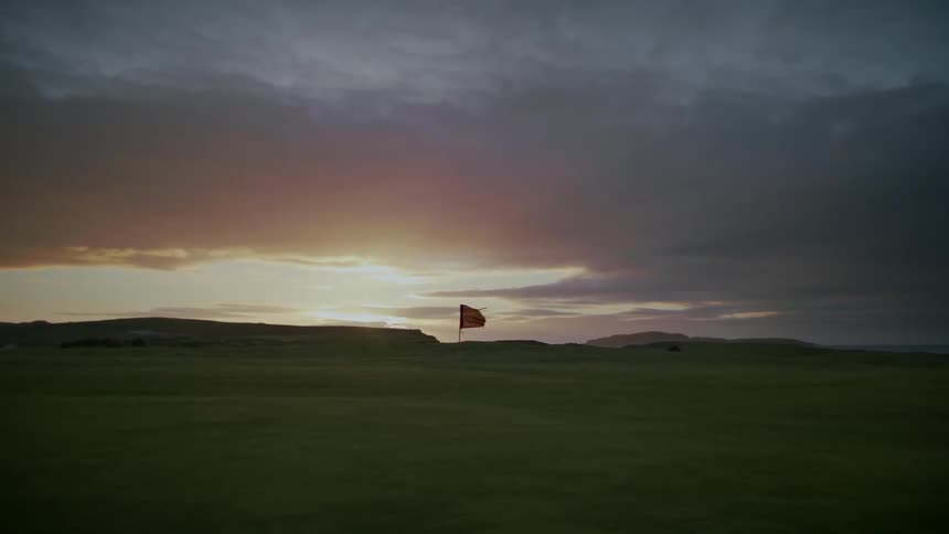Flag marking the hole on the green of a golf course blowing in the wind and a dark clouded sky with pink and white at dusk