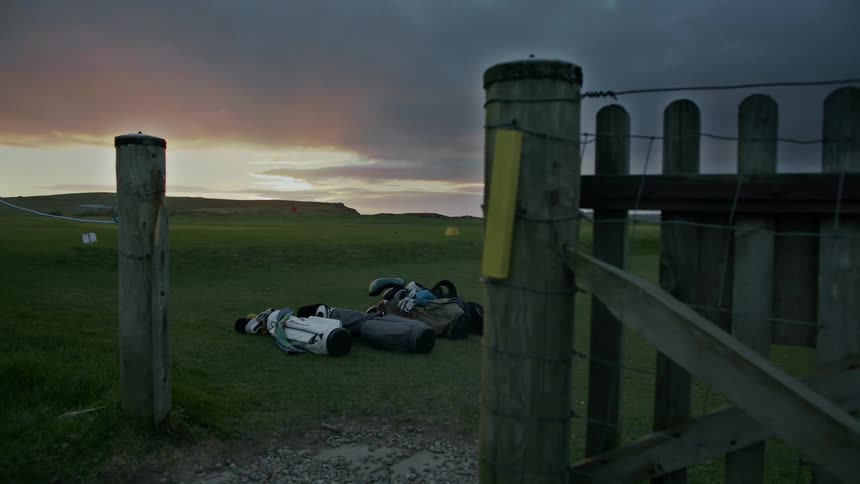Golf bags lay on the grass of a colf course near a wood gate with a purple and pink clouded sky at sunset