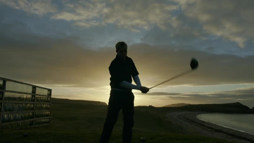 A silhouette of a man on a golf course swinging a golf club to hit a golf ball into the distance
