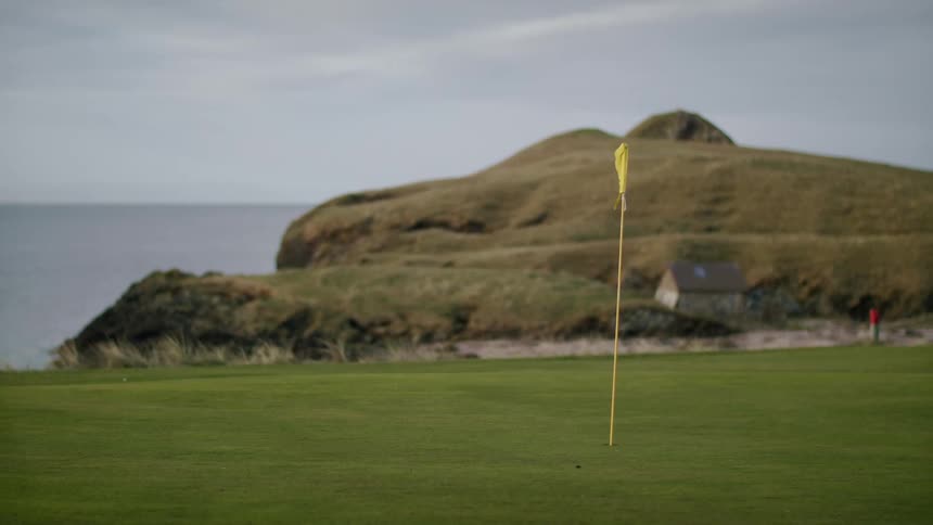 Yellow flag marks golf hole cup near hill overlooking ocean on an overcast day