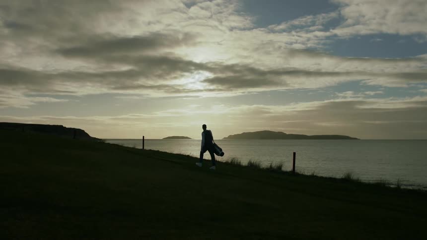 A man is carrying his golf bag by the shore