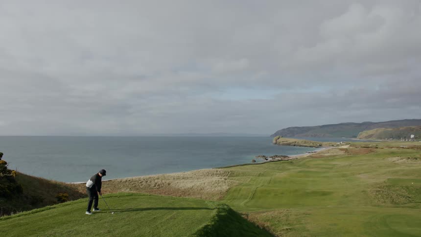 A man taking a golf swing on an open field near the sea on a sunny day