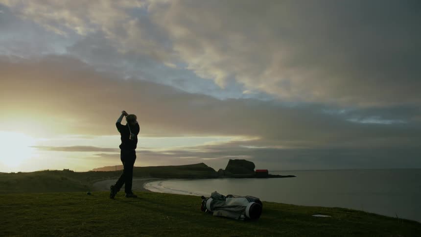 Man playing golf on a course by the sea under a cloudy sky with the sun on the horizon