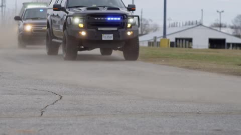 Unmarked police vehicles flash blue and red lights while driving along road past warehouses