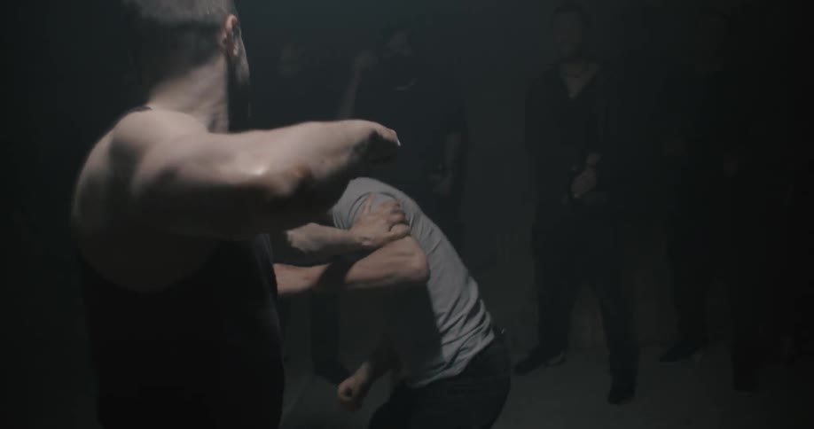 Two people are fist fighting while others watch in a dark room