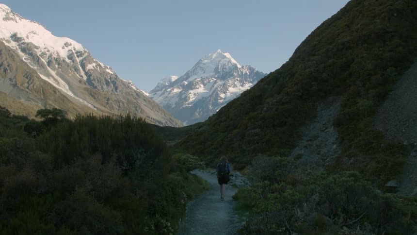 A woman is walking on a trail towards a snow covered mountain
