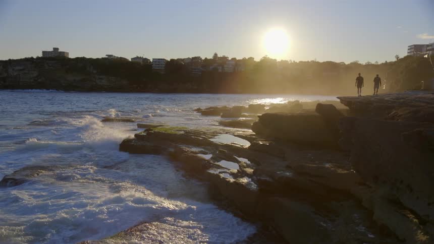 Two people walk on the rocks near the shore with heavy waves crashing