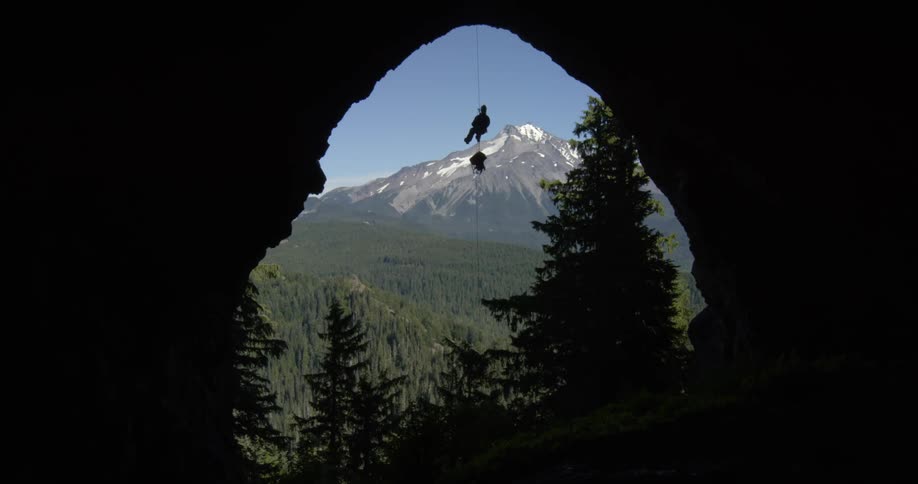 A rock climber slides down a hanging rope as viewed from a cavernous opening in the wilderness