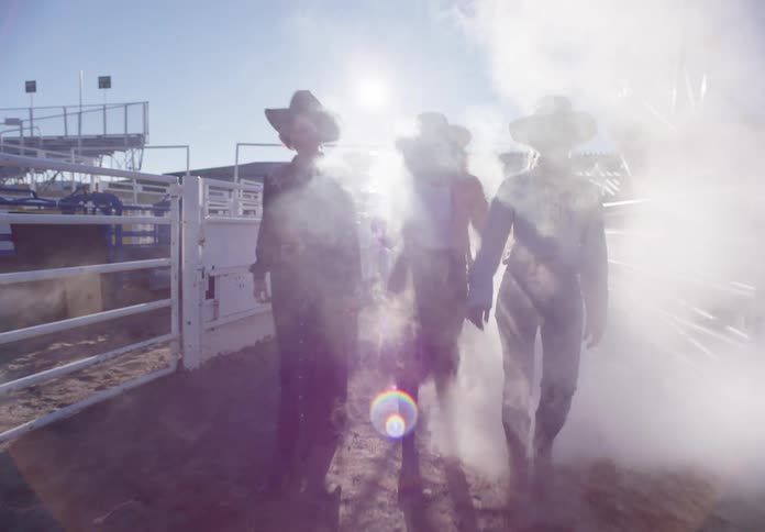 Three models dressed as cowgirls crossing through a curtain of smoke in a rodeo arena in daylight