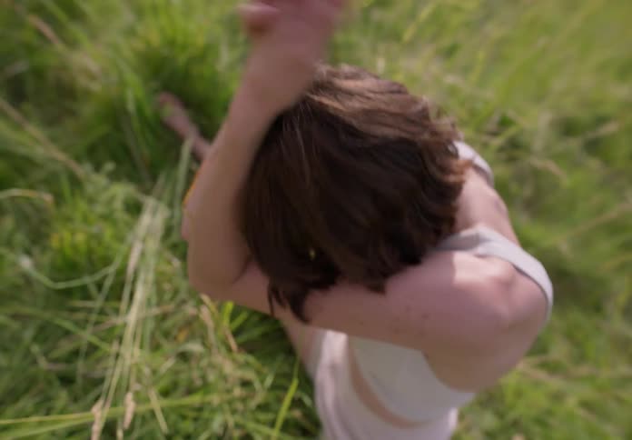 A woman sits in grass while moving around and putting grass in her hair