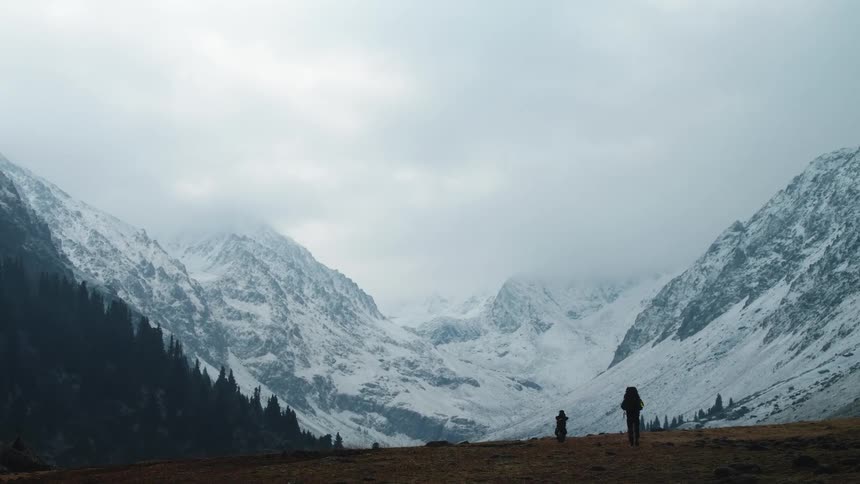Three people walking on a ground surrounded by a chain of snowcovered mountains on a cloudy day