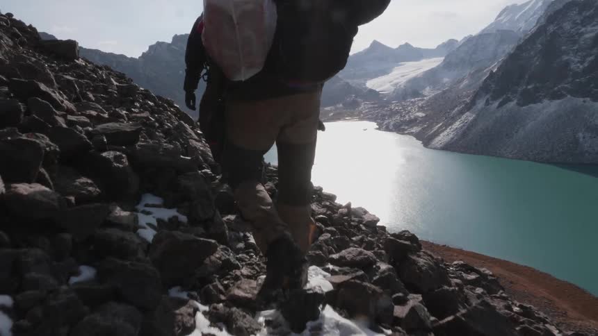Two people walking on a cliff with rocks next to a lake and a snowcovered mountain range in the background on a morning