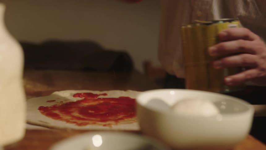 A man uses a wooden spoon to spread canned tomato sauce on a circle of pizza dough