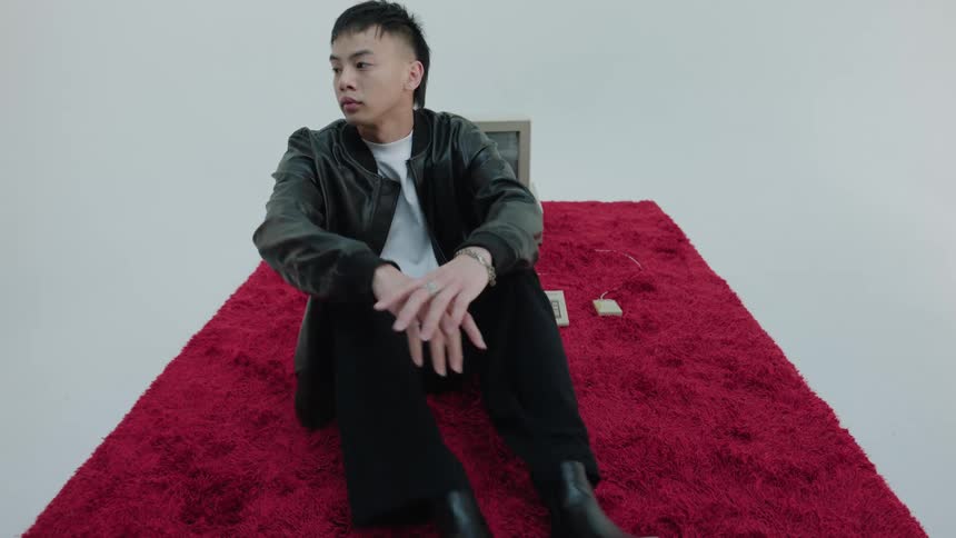 A man sitting on a red rug with an old computer on it turns around and uncrosses his legs facing the camera