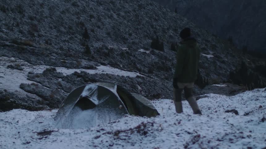 Person camping on snowy field at base of mountain approaches tent