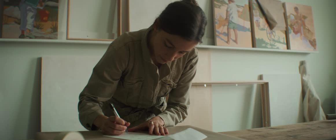 A woman is writing and a paper is on a table and there are paintings on the wall
