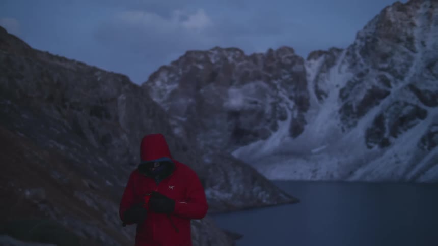 Man in a red jacket next to a blue lake with snowcapped mountains at the beginning of dusk