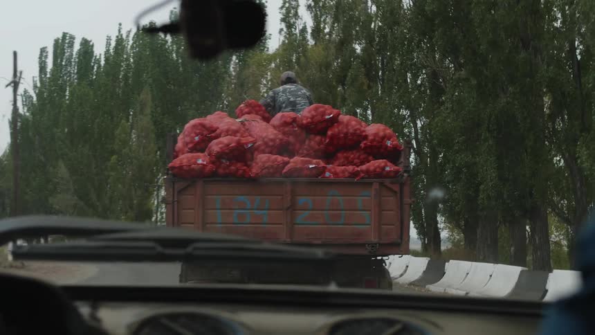 View of the car dashboard with a person on top of a moving truck bed carrying sacks of potatoes