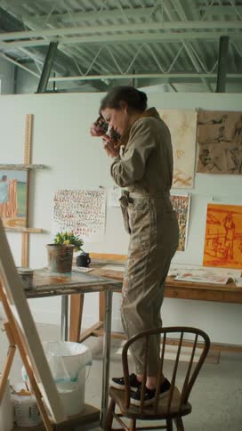 Artist stands on chair in art studio and takes picture of art lying on table