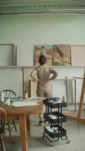 Woman hangs a finished painting on the wall in a studio, admires work