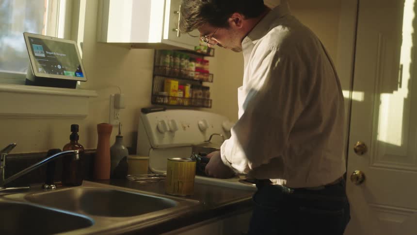 A man bent over a kitchen sink using a can opener to open a can