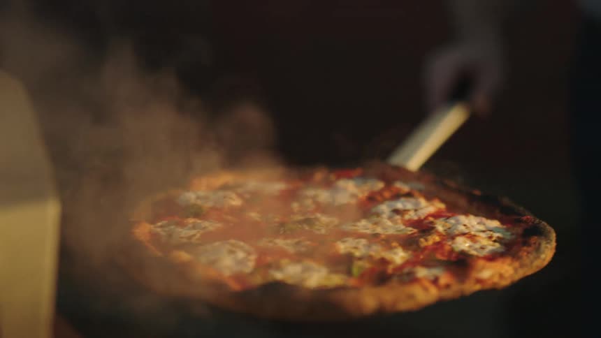 Hand of a man with a peel taking the pizza out of a woodfired oven