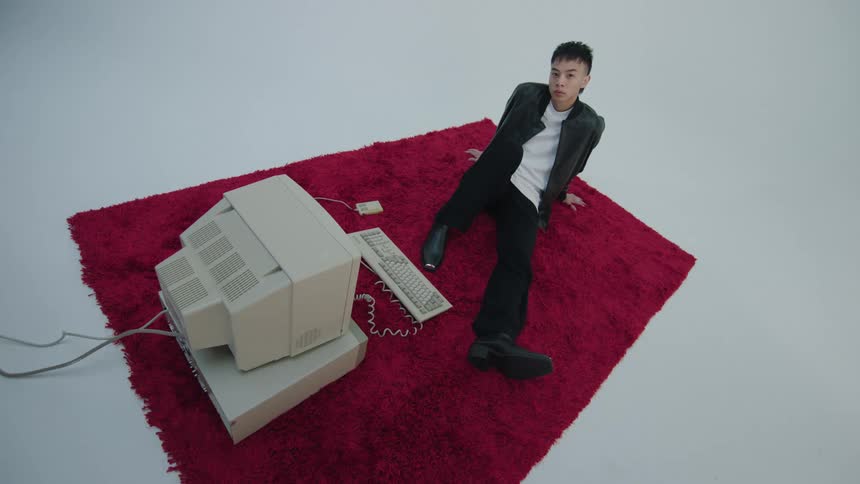 Boy wearing a black jacket pants and a white t shirt sits on a red rug watching old computer