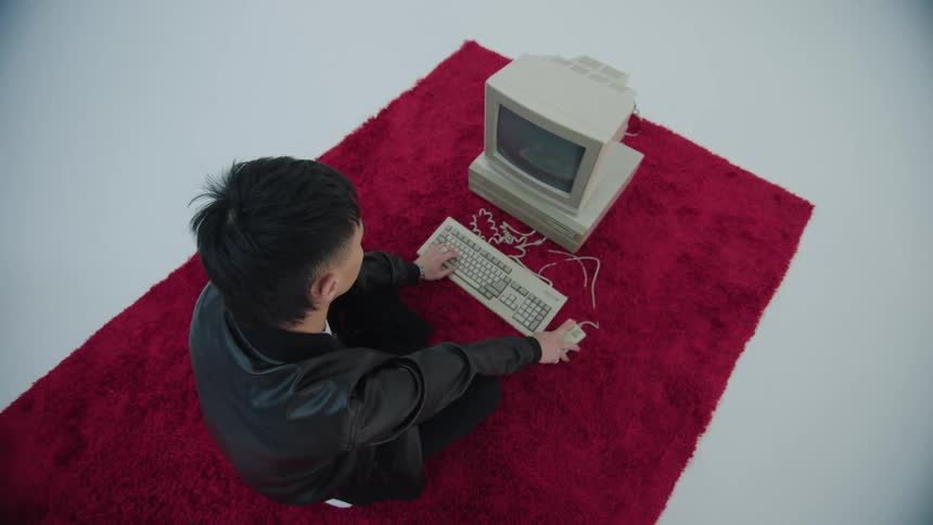 A young man typing on an old computer sitting on a rug