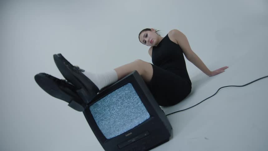 Woman in black dress poses with shoes propped up on television playing static