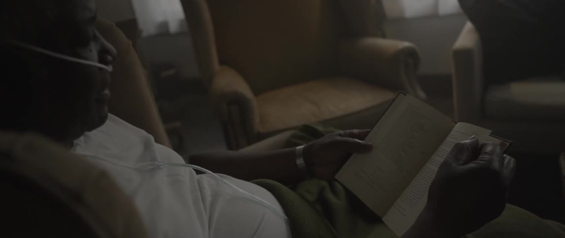 A man with a breathing tube sits in an armchair and reads