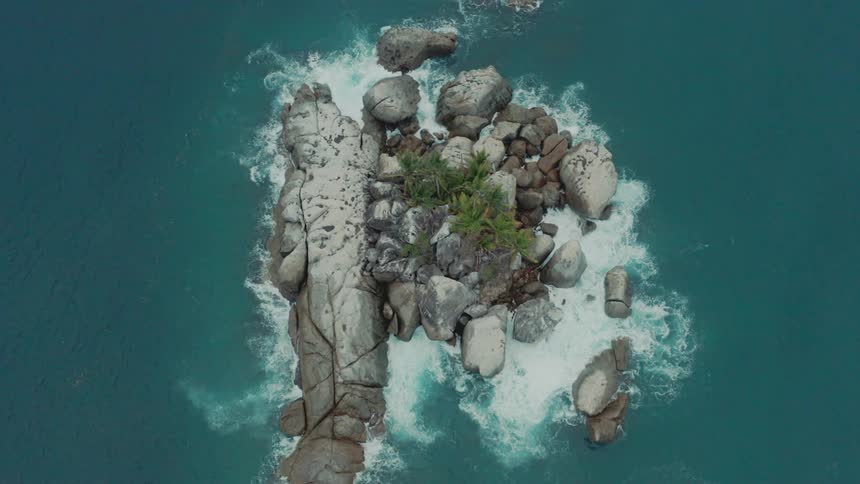 Aerial view of a small island in the midst of turquoise blue sea with waves crashing against rocks in slow motion