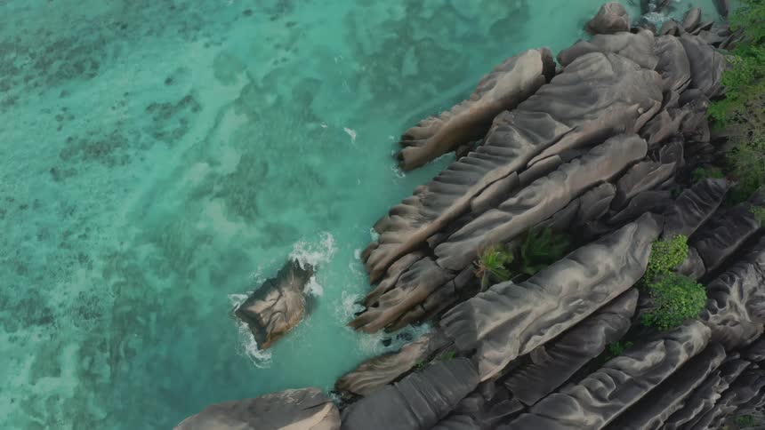 Aerial straight down perspective of waves crashing onto folded rocks next to a clear blue sea