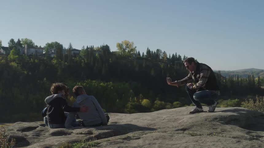 Mother sitting beside the children with the father taking a photo of them on a cliff in nature
