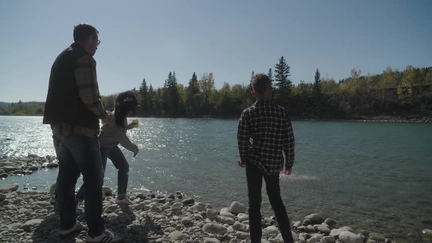 A family of 4 stand on the riverbank watching as one of them skips a rock over the water