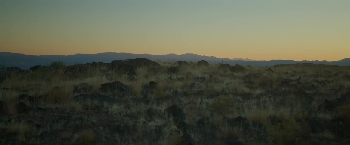 A cliff with grasses on top and a mountain view in a distance