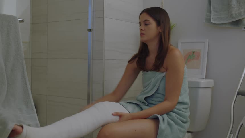 Woman sitting on the toilet in the bathroom drying her hands and supporting her leg which is in a cast and bandaged