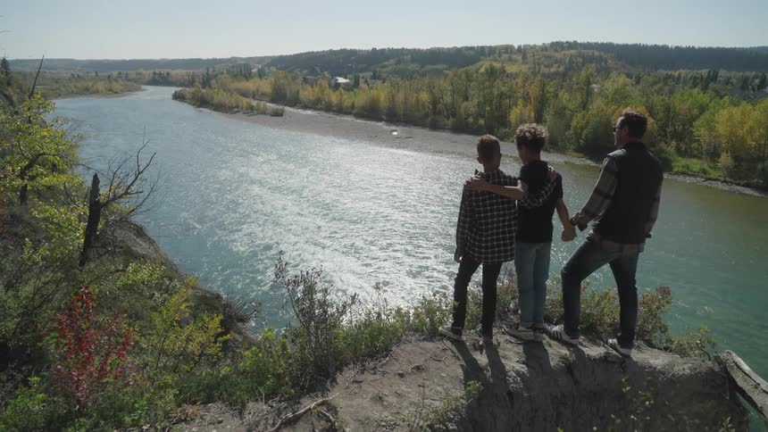 Two boys stand with a man holding hands close by each other on top of a hill over looking a river surrounding by trees below on a sunny day