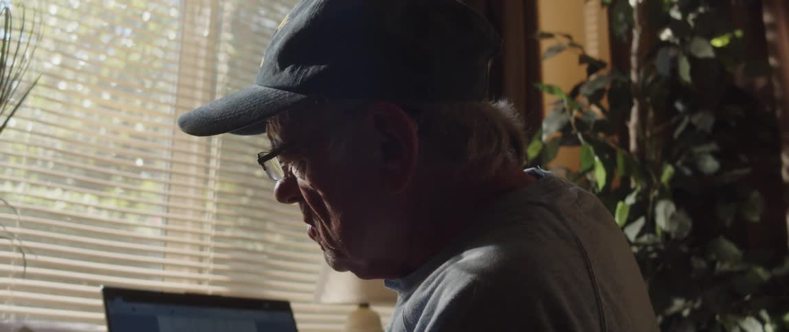 Man wearing baseball cap sits at desk laptop computer near window