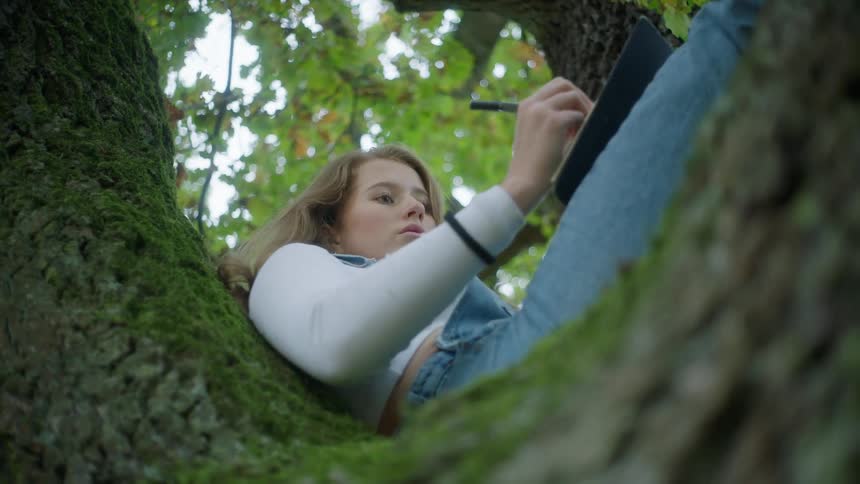 A young woman sitting up on a tree sketching on a notepad