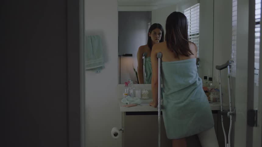 A woman with a broken leg with a towel wrapped around her standing in front of her bathroom mirror with the help of crutches