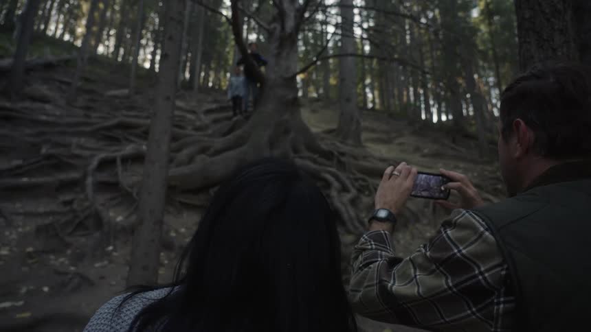 Two people are taking photos using a cell phone of a child in a huge tree with roots sticking out of the ground