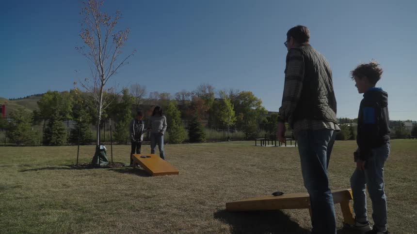 Family plays bean bag toss in park near hills on sunny day