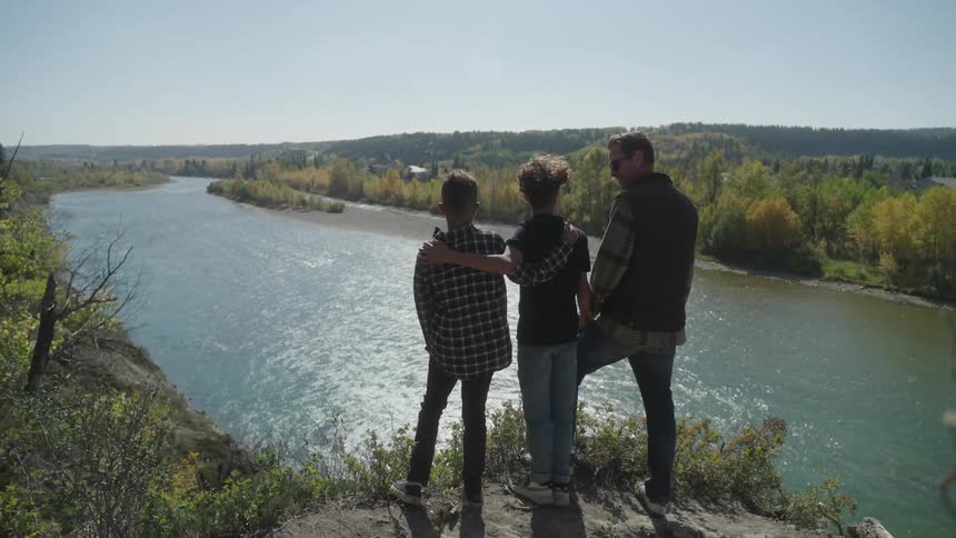 A father and his two teenage sons stand on a hill looking out towards a river