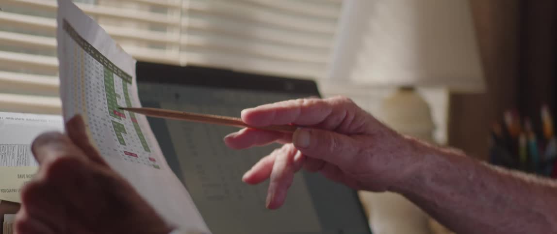 Elderly person facing a laptop and holding a paper while analyzing data