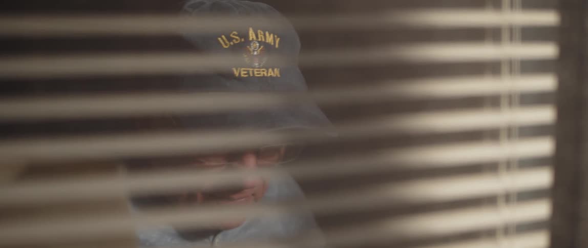 Window blinds obscure face of elderly man donning us army baseball cap