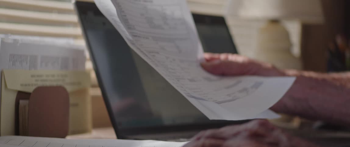 A man is shuffling through paperwork while sitting at a desk with a laptop in the daytime