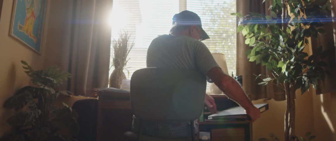 Elderly person sitting at a desk with their back to the camera reaching for a notebook